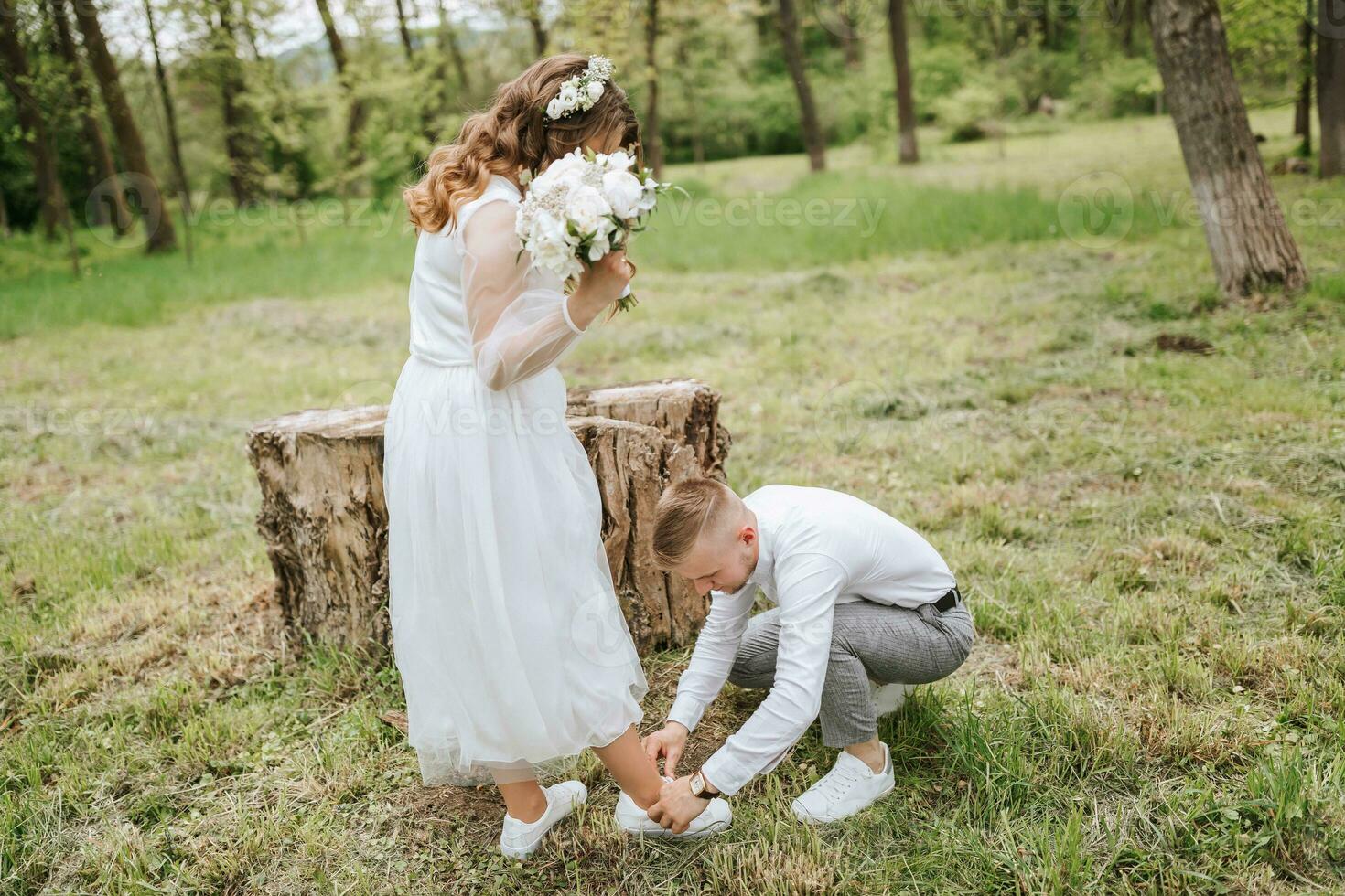 bridal walk in the forest. The groom is dressed in a white shirt and gray pants, helping the bride to tie her shoelaces photo