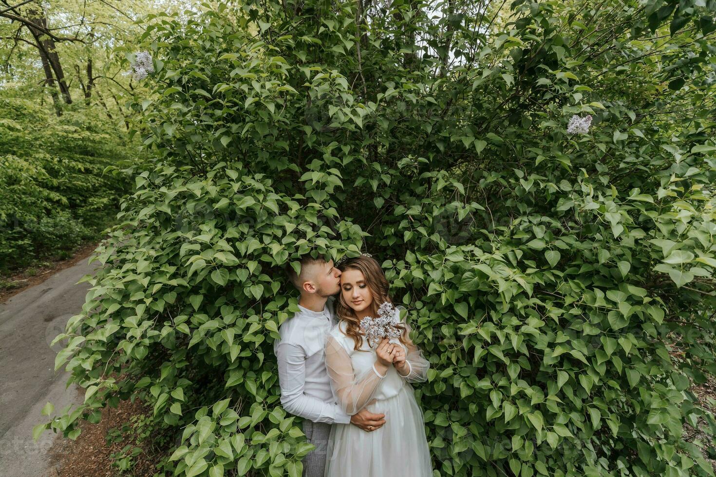 bride and groom stand on a background of green leaves. The groom kisses the bride on the temple photo