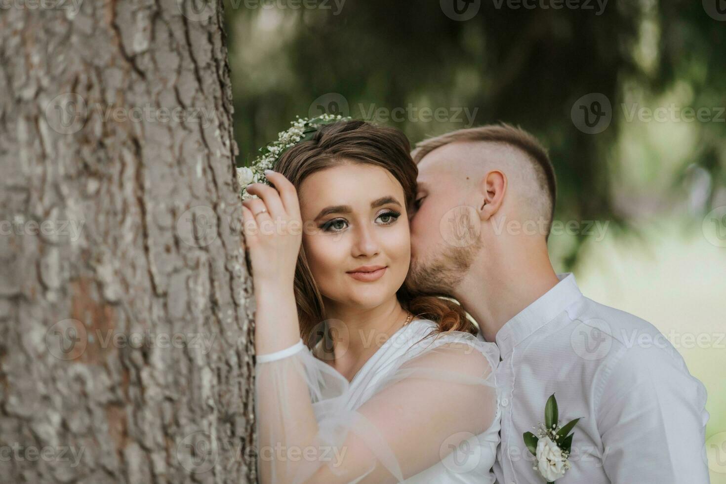 retrato de el novia y novio. un novia con largo pelo y un guirnalda en su cabeza cerca un alto árbol, desde detrás el novio abrazos su y Besos su en el mejilla. foto