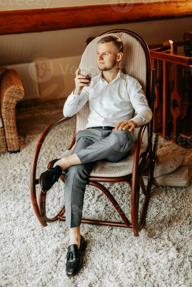 un joven empresario en un hotel habitación se sienta en un silla y bebidas su whisky desde un vaso. descanso en maravilloso apartamentos del novio Mañana a el hotel foto