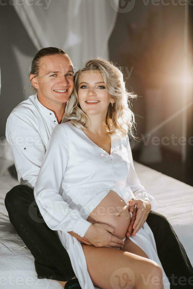 studio portrait of beautiful pregnant young woman in white dress sitting on bed with man, hugging and happy photo