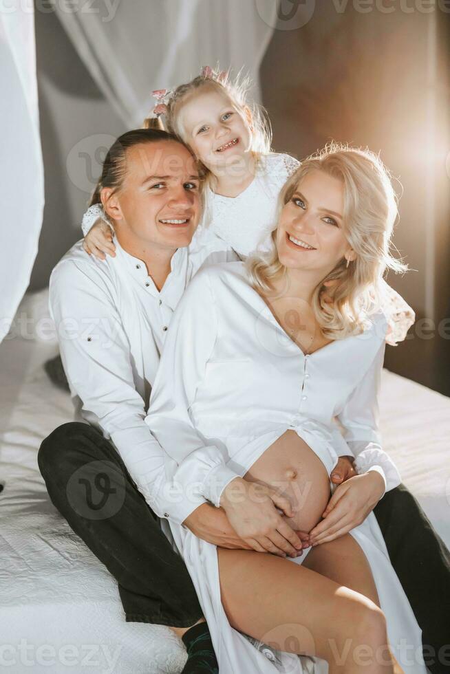 studio portrait of beautiful pregnant young woman in white dress sitting on bed with man, hugging and happy photo
