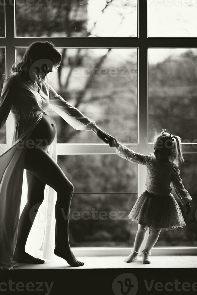 Studio portrait of a beautiful pregnant young woman near the window with her daughter. Black and white photo