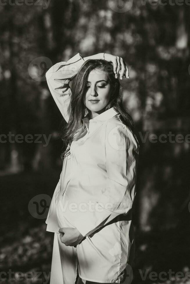 A pregnant woman in a white dress on a dark background poses in the park. Happy and carefree pregnancy. Autumn park. Happy woman enjoying her pregnancy. photo