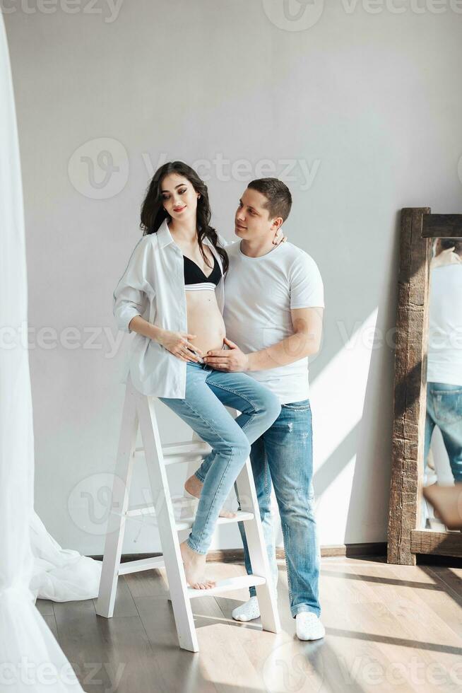 modern beautiful pregnant woman in jeans and shirt with man hugging her tummy with hands in beautiful sunlight. Concept of pregnancy, motherhood, preparation and waiting. photo