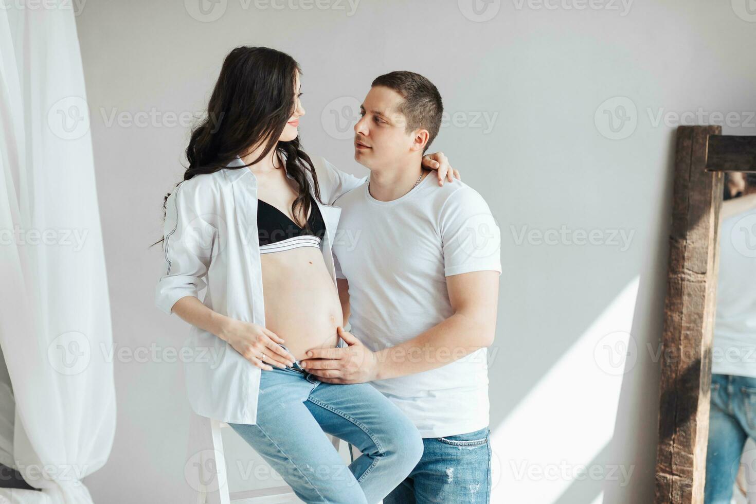 moderno hermosa embarazada mujer en pantalones y camisa con hombre abrazando su barriguita con manos en hermosa luz de sol. concepto de el embarazo, maternidad, preparación y espera. foto