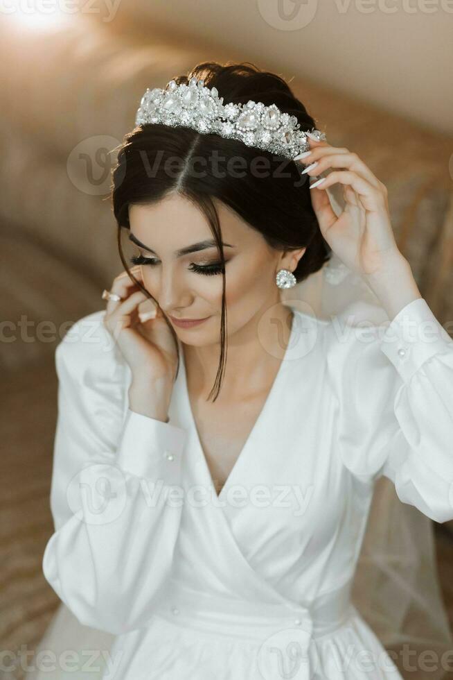 portrait of a beautiful girl in a robe with a crown on her head. Fashion, glamour, concepts. The morning of the bride, the bride in a robe is sitting on the couch. photo