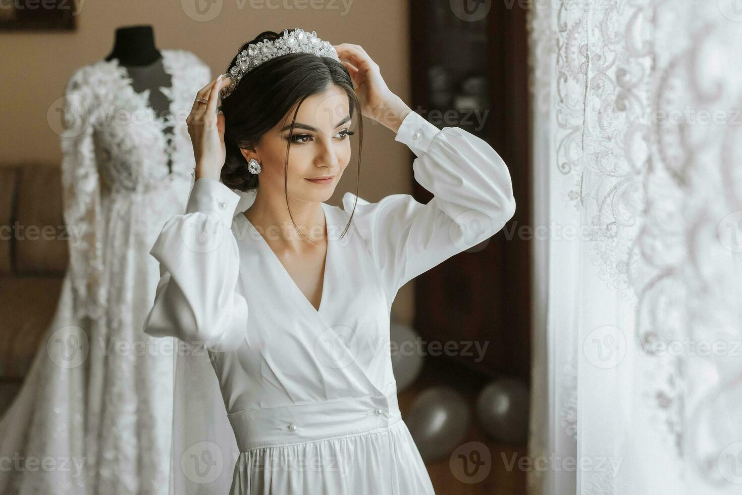 portrait of a beautiful girl in a robe with a crown on her head. Fashion, glamour, concepts. The morning of the bride, the bride in a robe stands near a mannequin with her wedding dress photo