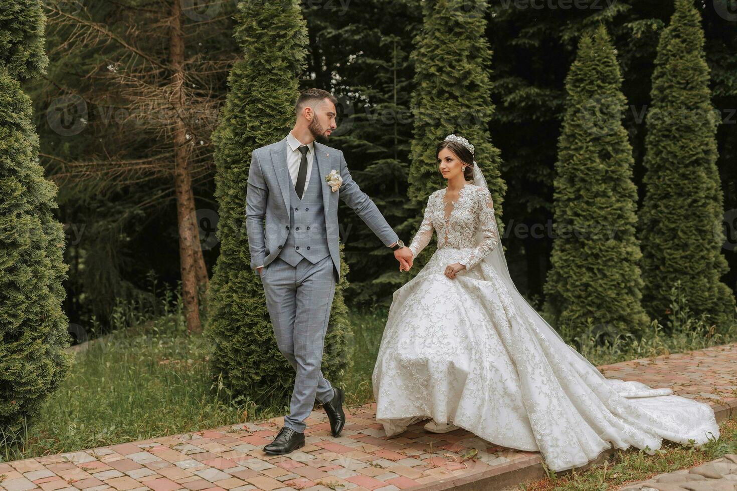 European wedding couple walking in the park. The bride in a beautiful dress with sleeves and a crown on her head. Groom with a black beard of Caucasian appearance in a classic suit photo