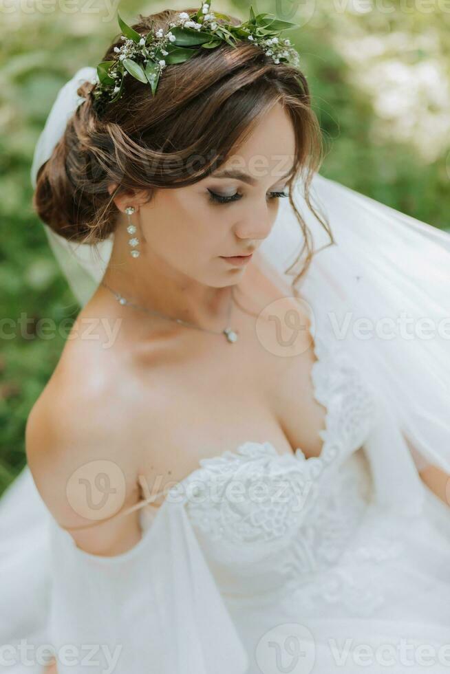 un hermosa novia en un blanco vestir con un guirnalda de Fresco flores es sentado en el verde césped foto