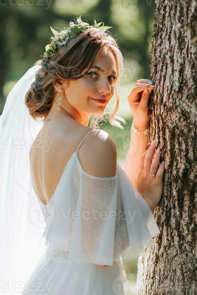 Portrait of a beautiful cute bride girl in an elegant white dress posing near a tree in the forest on a sunny summer day photo