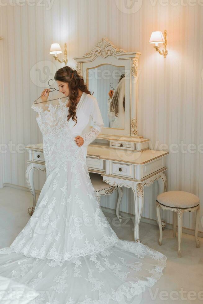 A beautiful young bride in a white petticoat is standing and holding a wedding dress in her hands. Final preparations for the wedding. The bride is waiting for the groom. Morning, bride. photo