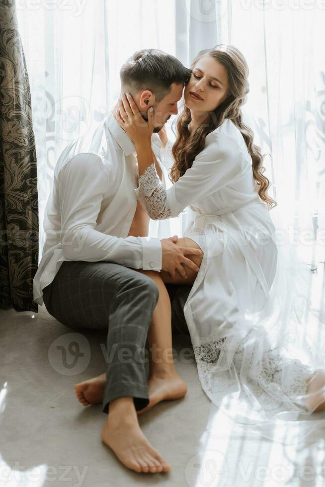 Beautiful, sexy bride in a white robe, groom in a white shirt hugs and kisses the bride against the background of a window in a hotel room. Wedding portrait of newlyweds in love. Vertical photo. photo