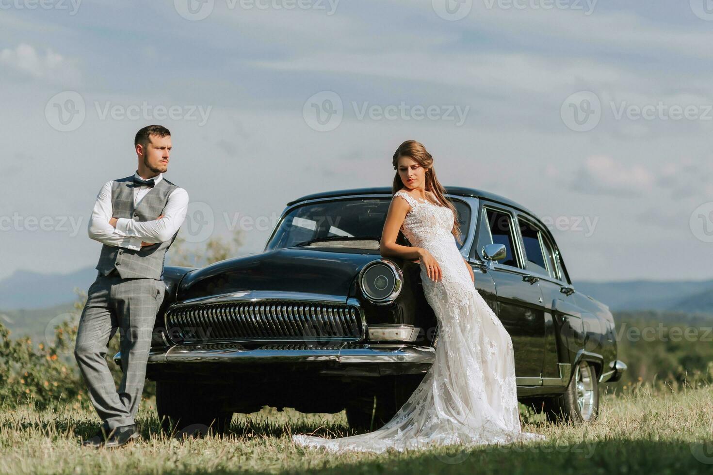 Front view of a married bride and groom wearing festive clothes standing against a black retro car on their wedding day photo