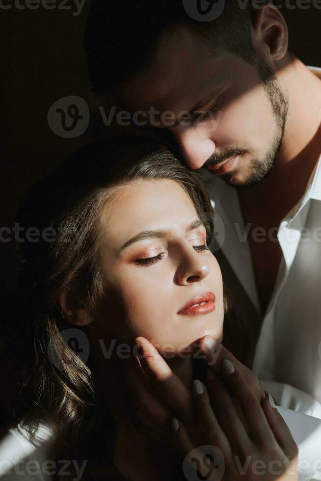 Beautiful, sexy bride in a white robe, groom in a white shirt hugs the bride in a hotel room. Wedding portrait of newlyweds in love. photo