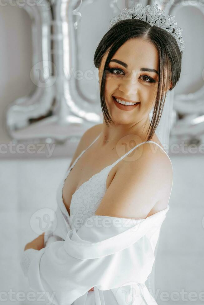 A beautiful bride girl in a white robe, veil and tiara on her head, the sun shines on her from the window. Happy young girl under the glare of sunlight. wedding concept photo