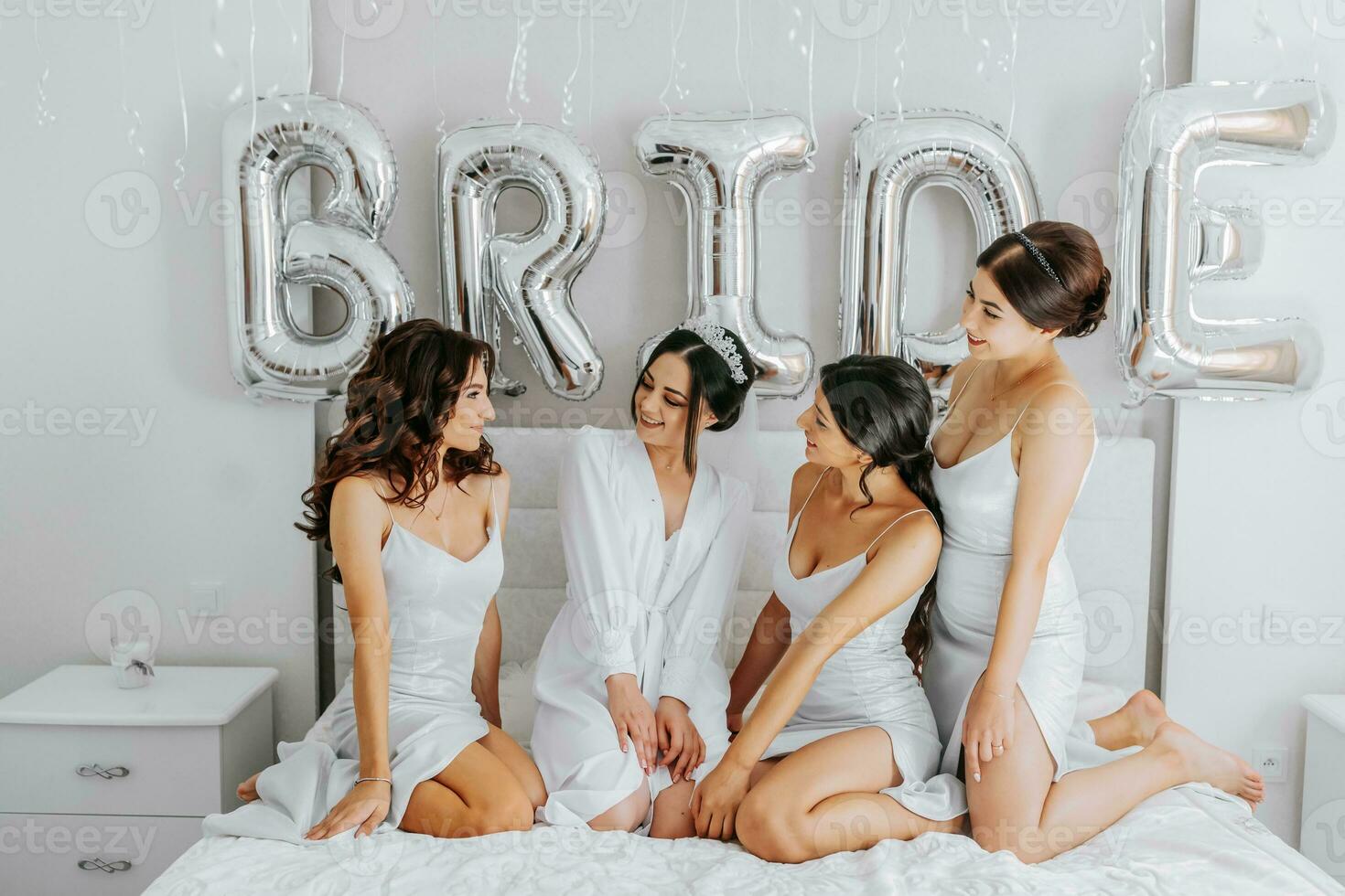 Young bridesmaids in white silk dresses on a bed in the bride's room. Beautiful women celebrating bachelorette party sitting on bed. photo