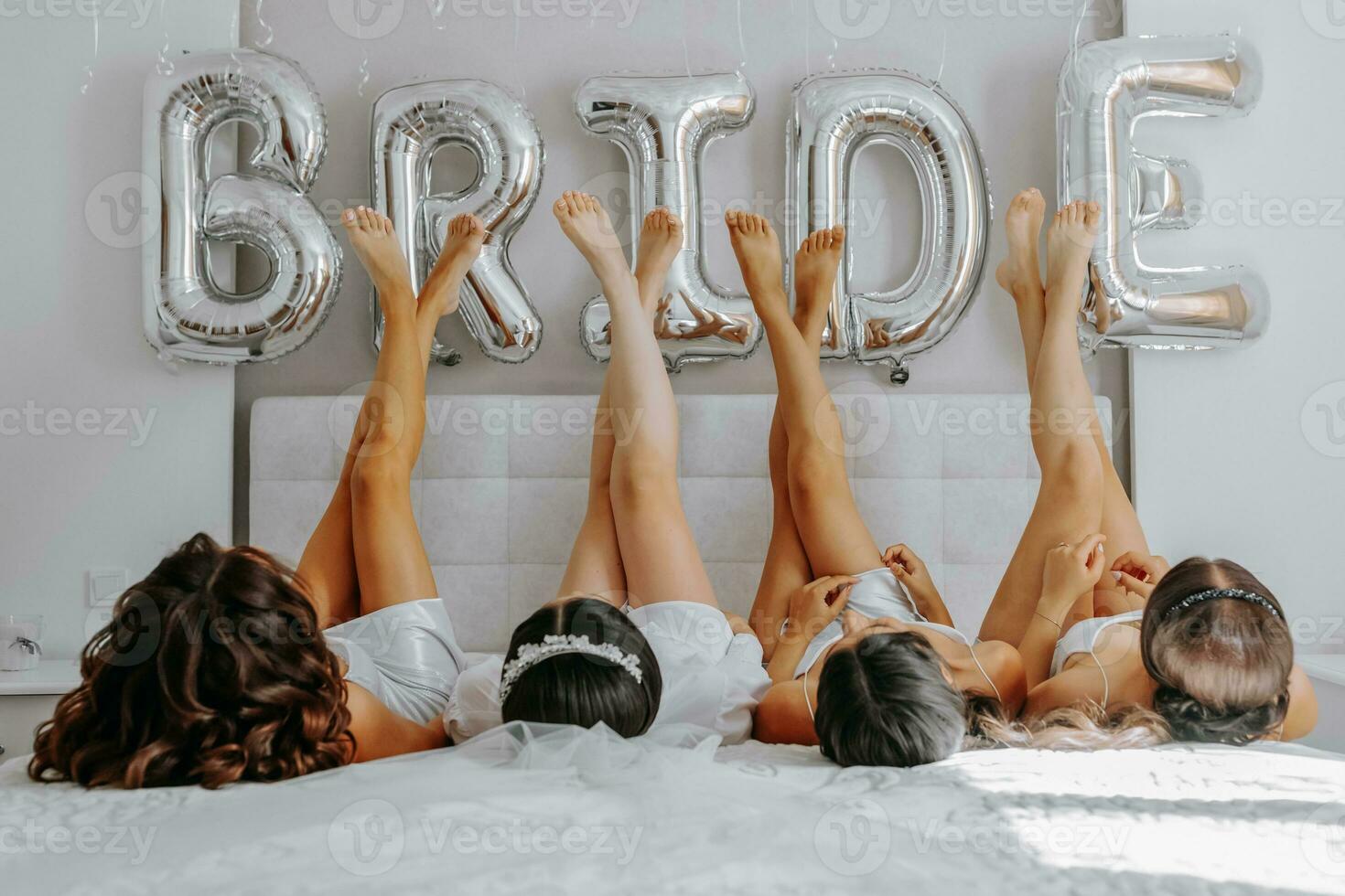 joven damas de honor en blanco seda vestidos en un cama en el de la novia habitación. hermosa mujer celebrando despedida de soltera fiesta sentado en cama. foto