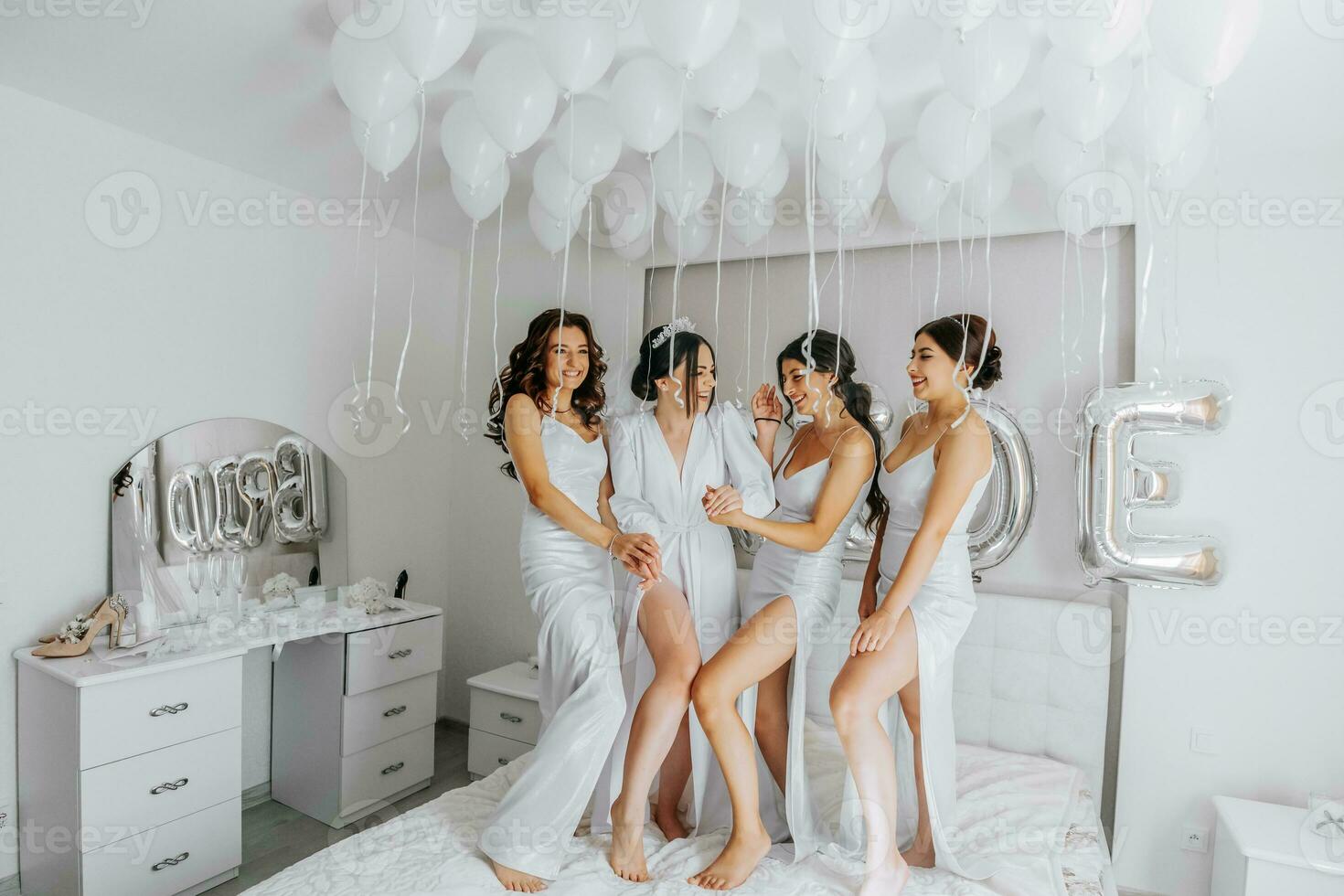 Young bridesmaids in white silk dresses on a bed in the bride's room. Beautiful women celebrating bachelorette party sitting on bed. photo