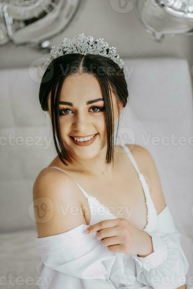 un hermosa novia niña en un blanco túnica, velo y tiara en su cabeza, el Dom brilla en su desde el ventana. contento joven niña debajo el destello de luz de sol. Boda concepto foto