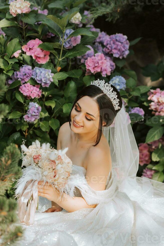 young beautiful bride in wedding dress with open shoulders and crown on her head sitting near hydrangea flowers, fashion photo taken under harsh sunlight