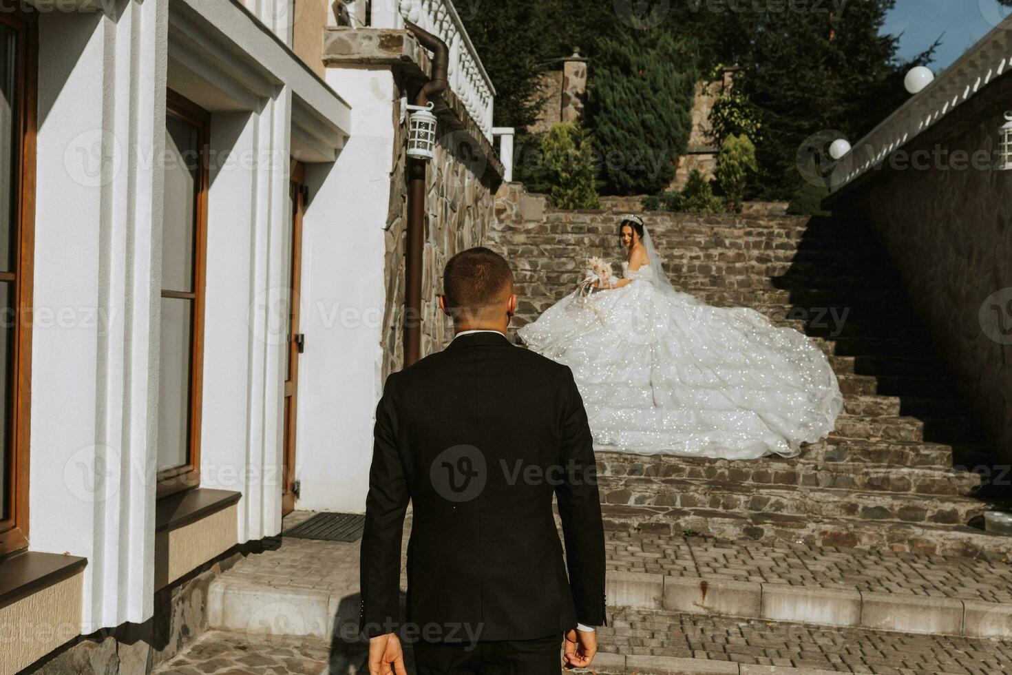 young beautiful bride in a wedding dress with open shoulders and a crown on her head is lying on stone stairs, fashion shot under bright sunlight. The groom goes to the bride photo