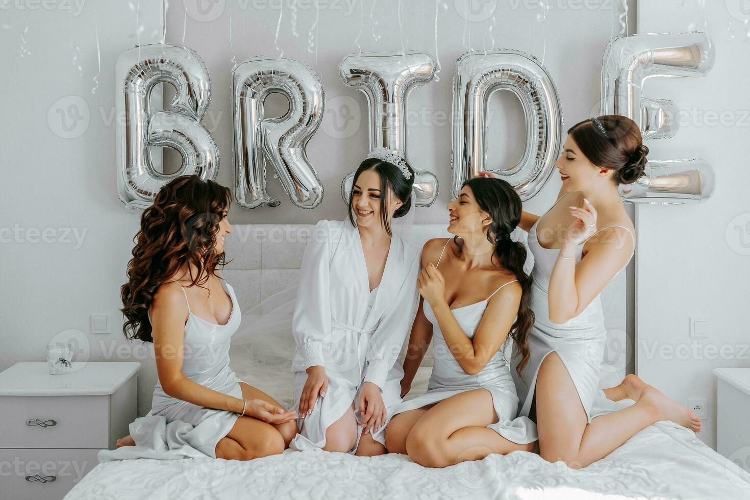 Young bridesmaids in white silk dresses on a bed in the bride's room. Beautiful women celebrating bachelorette party sitting on bed. photo