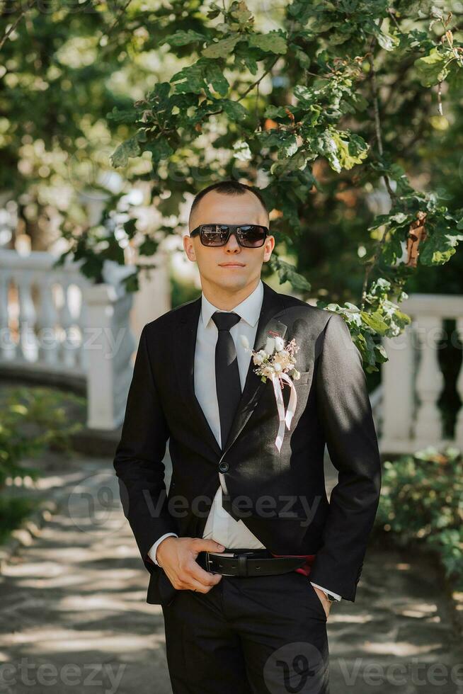 outdoor portrait of a young stylish groom in a classic black suit, white shirt and tie, with black glasses on his head photo