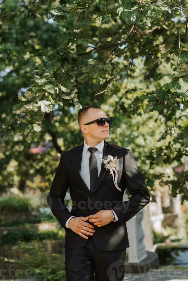 outdoor portrait of a young stylish groom in a classic black suit, white shirt and tie, with black glasses on his head photo