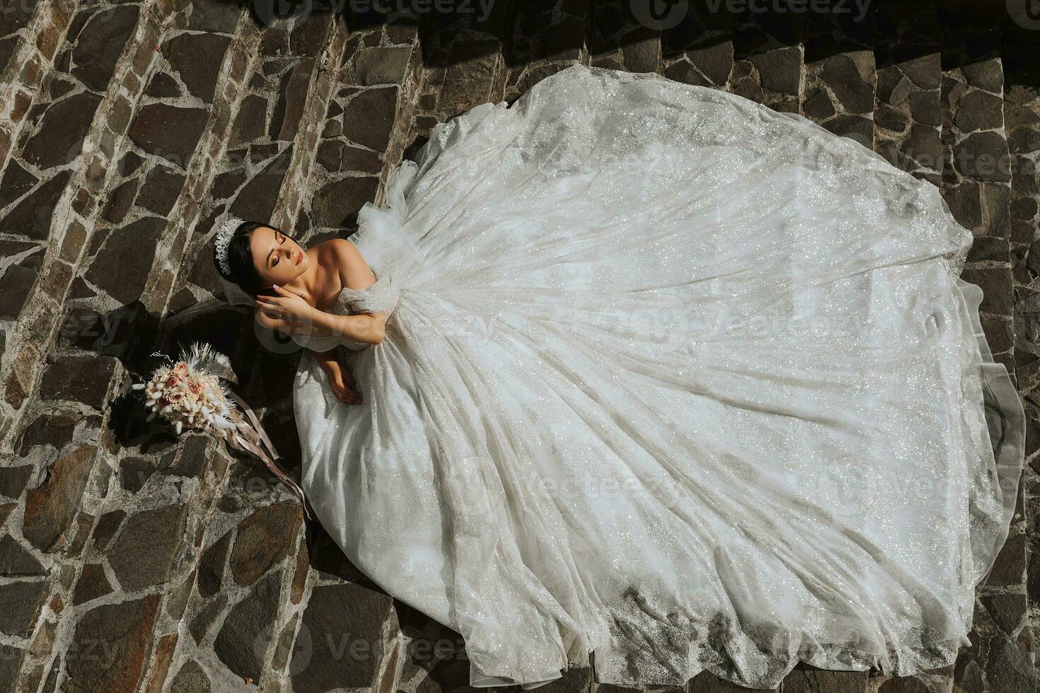 young beautiful bride in a wedding dress with open shoulders and a crown on her head is lying on stone stairs, fashion shot under harsh sunlight. The photo was taken from above