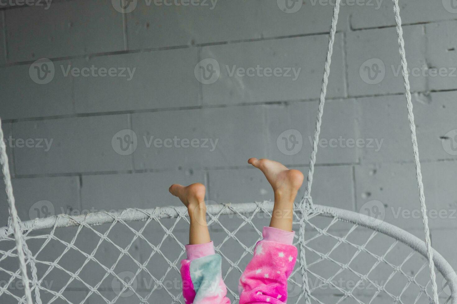 Happy little girl on the porch sitting on a wicker swing and playing in pink pajamas, legs up, close-up photo
