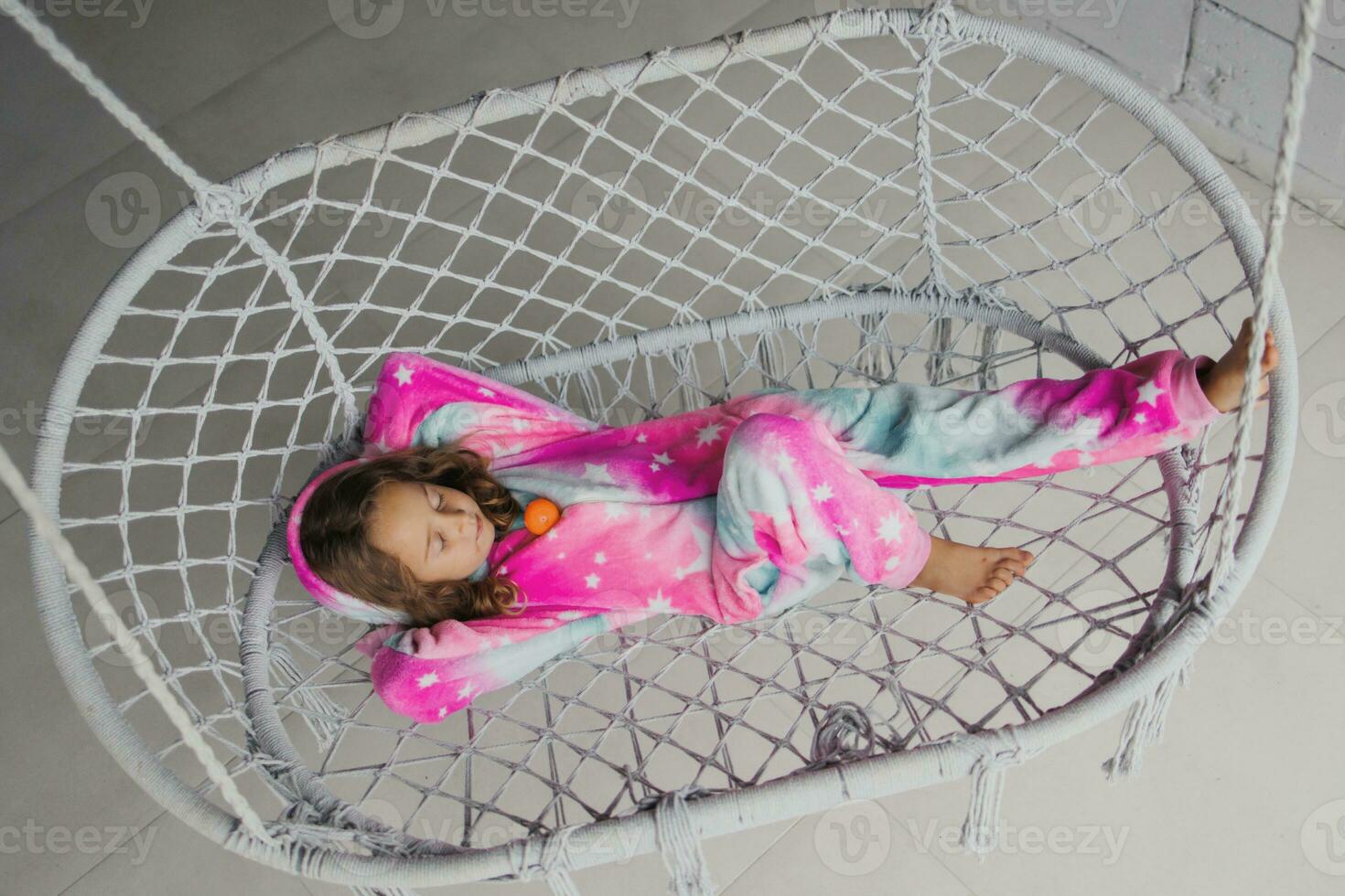 Happy little girl on the porch lying on a wicker swing and playing in pink pajamas with her little toy photo