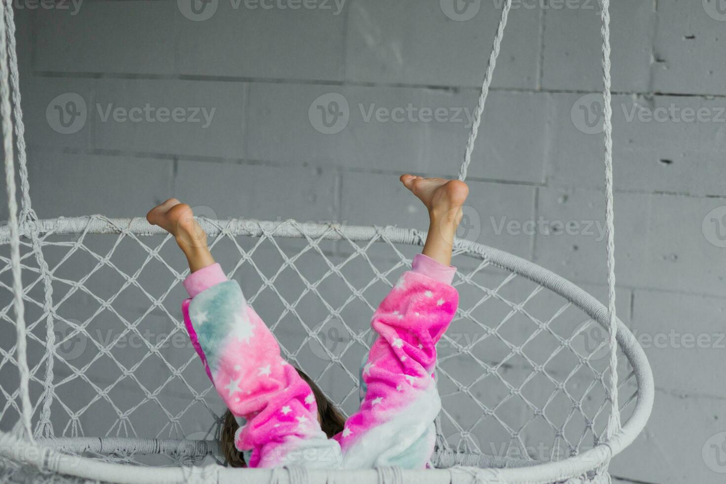 contento pequeño niña en el veranda sentado en un mimbre columpio y jugando en rosado pijama, levanta su piernas a el parte superior foto