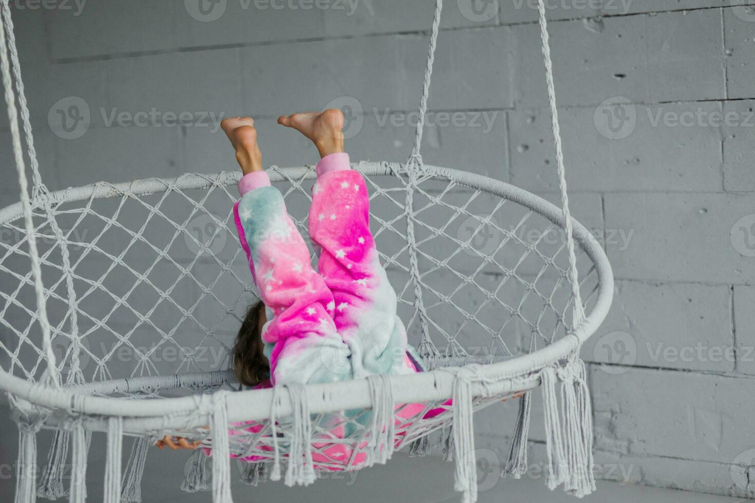 contento pequeño niña en el veranda sentado en un mimbre columpio y jugando en rosado pijama, levanta su piernas a el parte superior foto
