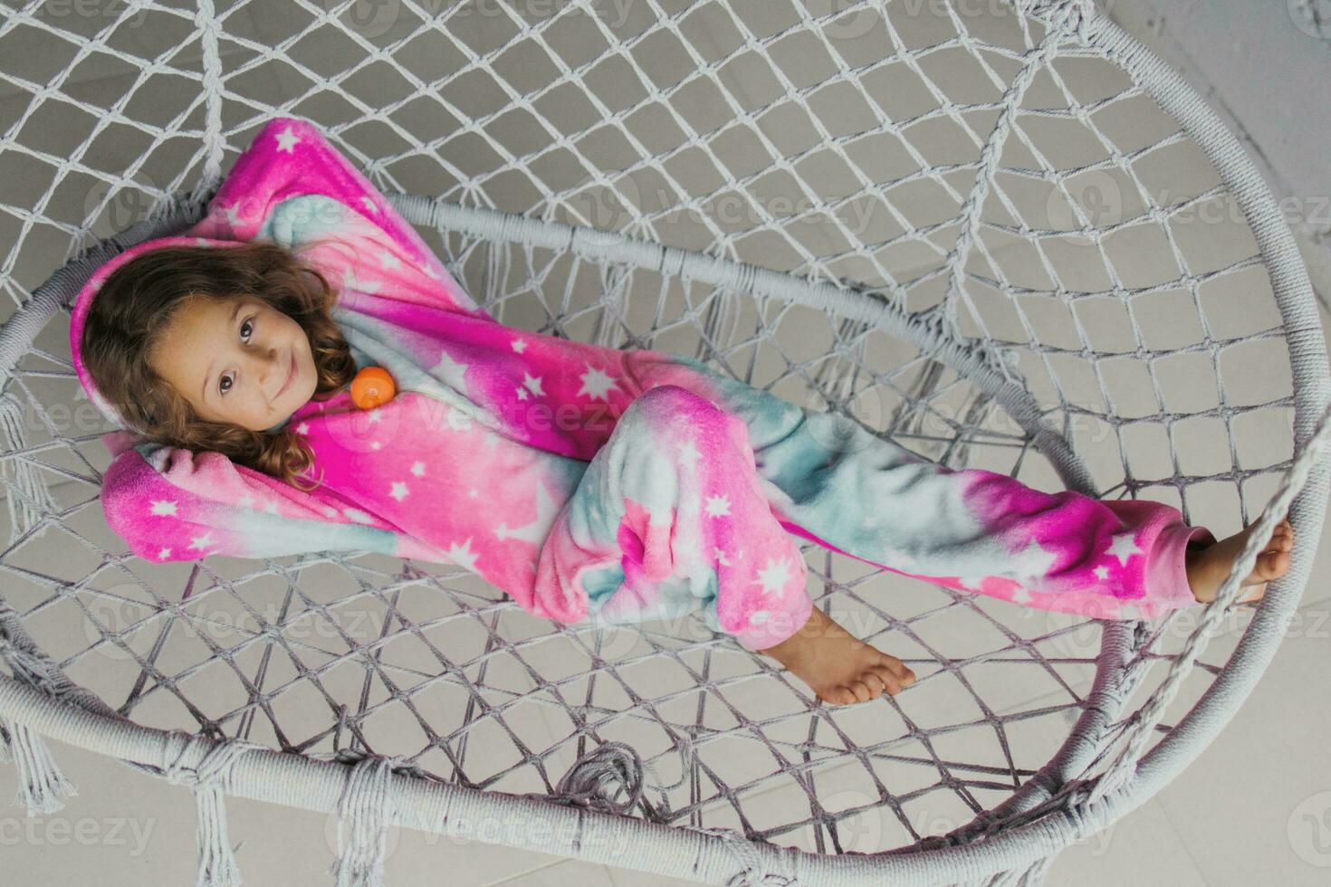 Happy little girl on the porch lying on a wicker swing and playing in pink pajamas with her little toy photo