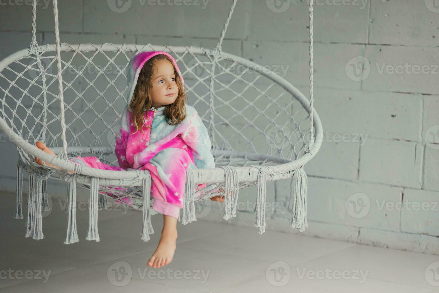 Portrait of happy little girl on porch sitting on wicker swing and playing in pink pajamas photo