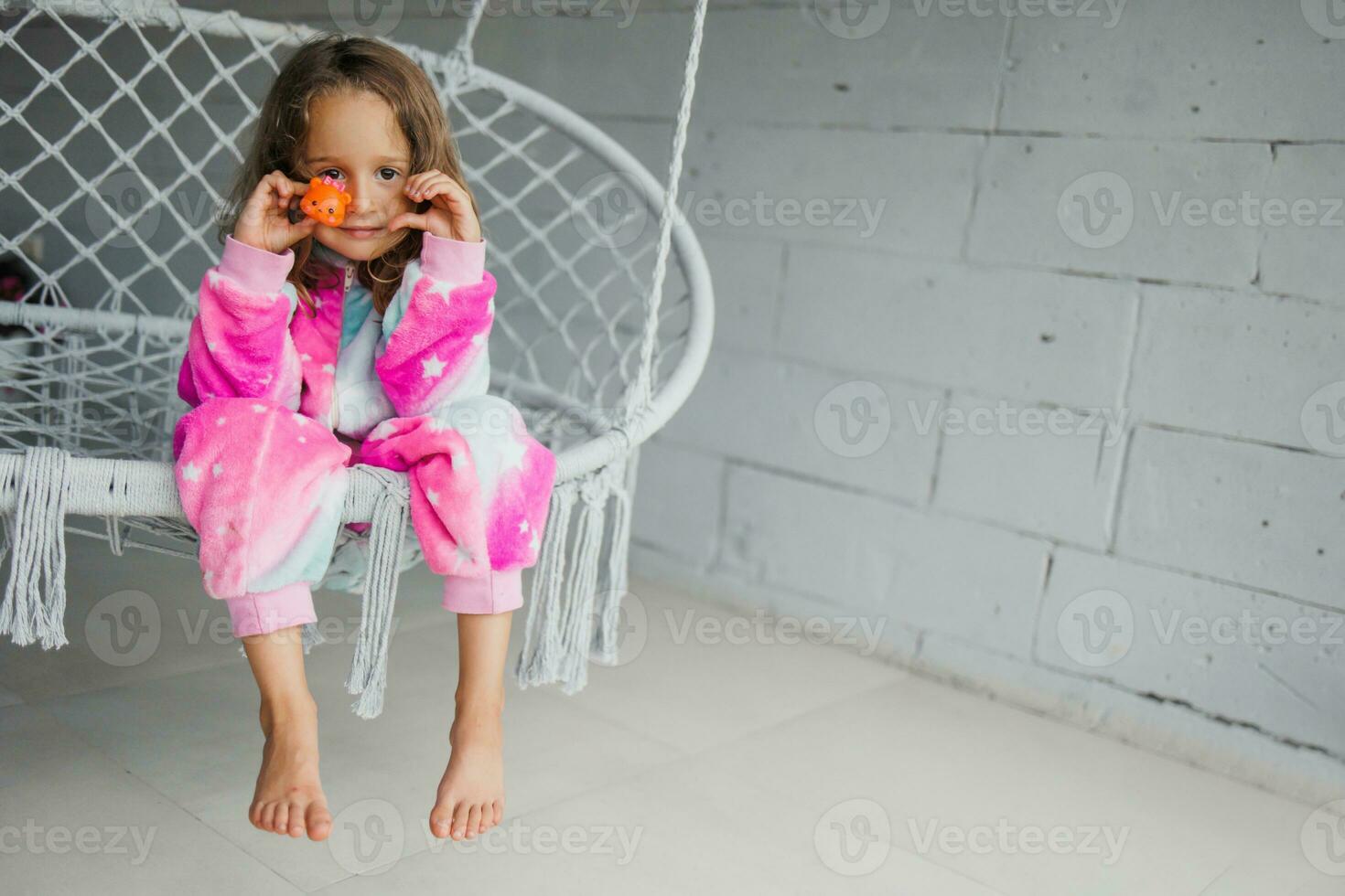 retrato de un contento pequeño niña en rosado pijama en el veranda, acostado en un mimbre columpio y jugando con su pequeño juguete foto