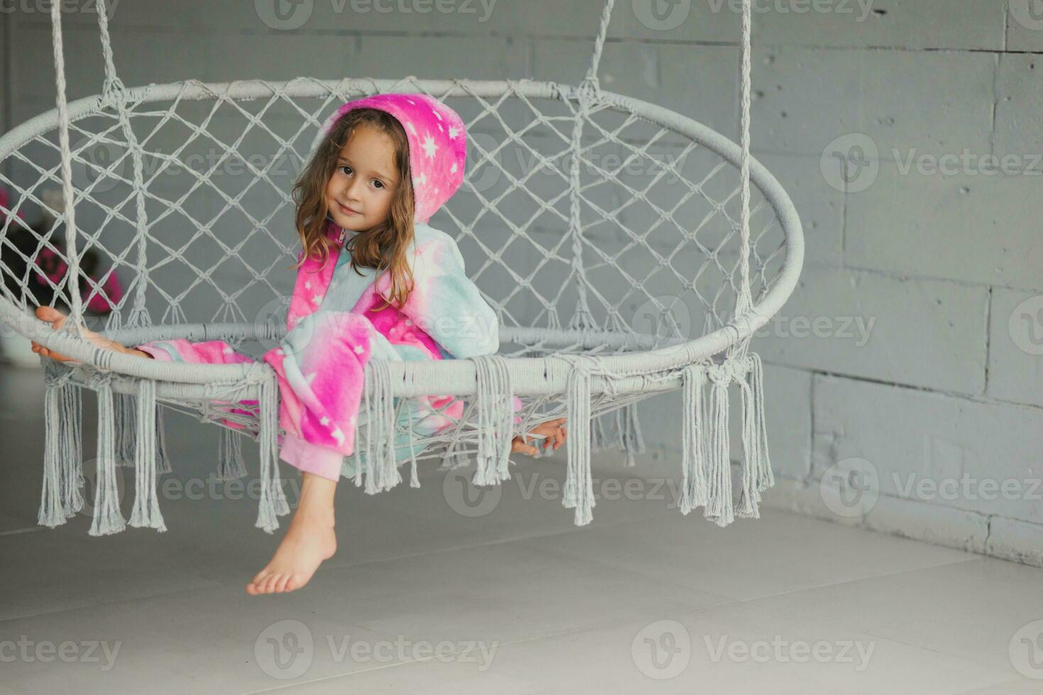 retrato de contento pequeño niña en porche sentado en mimbre columpio y jugando en rosado pijama foto