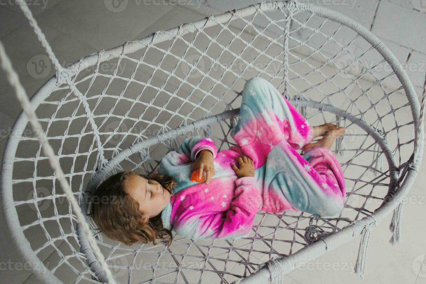 contento pequeño niña en el porche acostado en un mimbre columpio y jugando en rosado pijama con su pequeño juguete foto