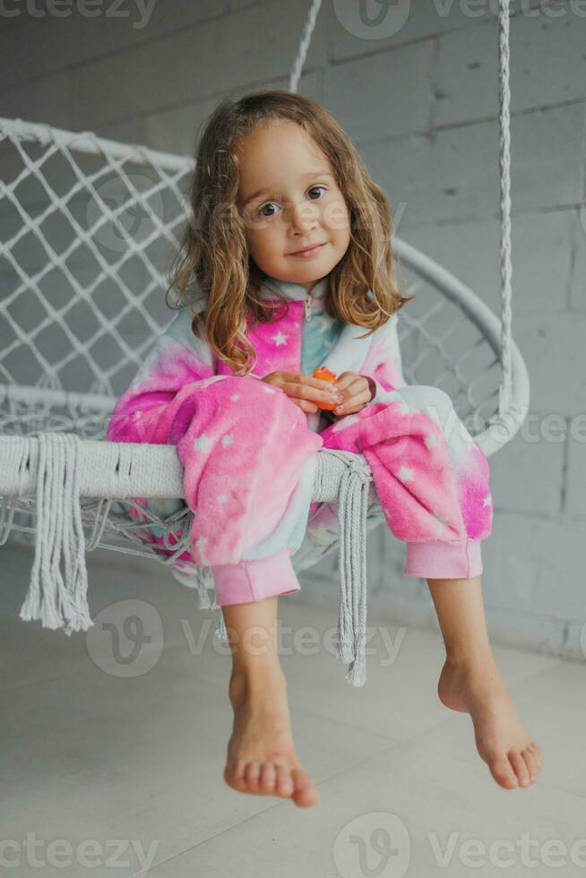 retrato de un contento pequeño niña en rosado pijama en el veranda, acostado en un mimbre columpio y jugando con su pequeño juguete foto