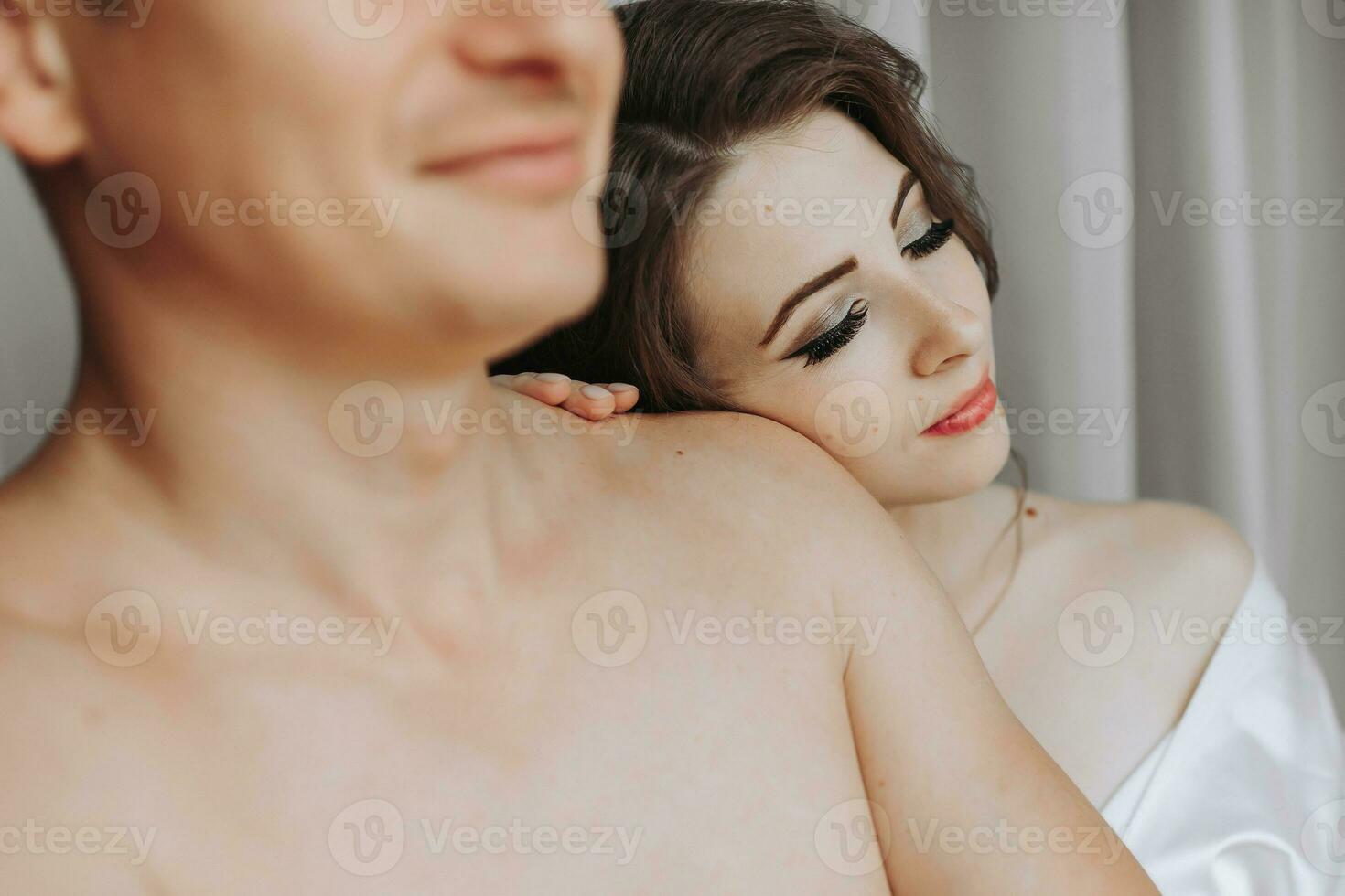 Beautiful, sexy bride in a white robe, bare-chested groom hugs and kisses the bride against the background of a window in a hotel room. Wedding portrait of newlyweds in love. photo