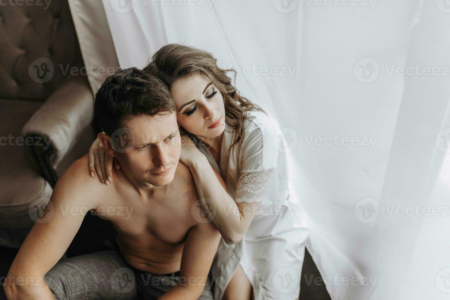 Beautiful, sexy bride in a white robe, bare-chested groom hugs and kisses the bride against the background of a window in a hotel room. Wedding portrait of newlyweds in love. photo