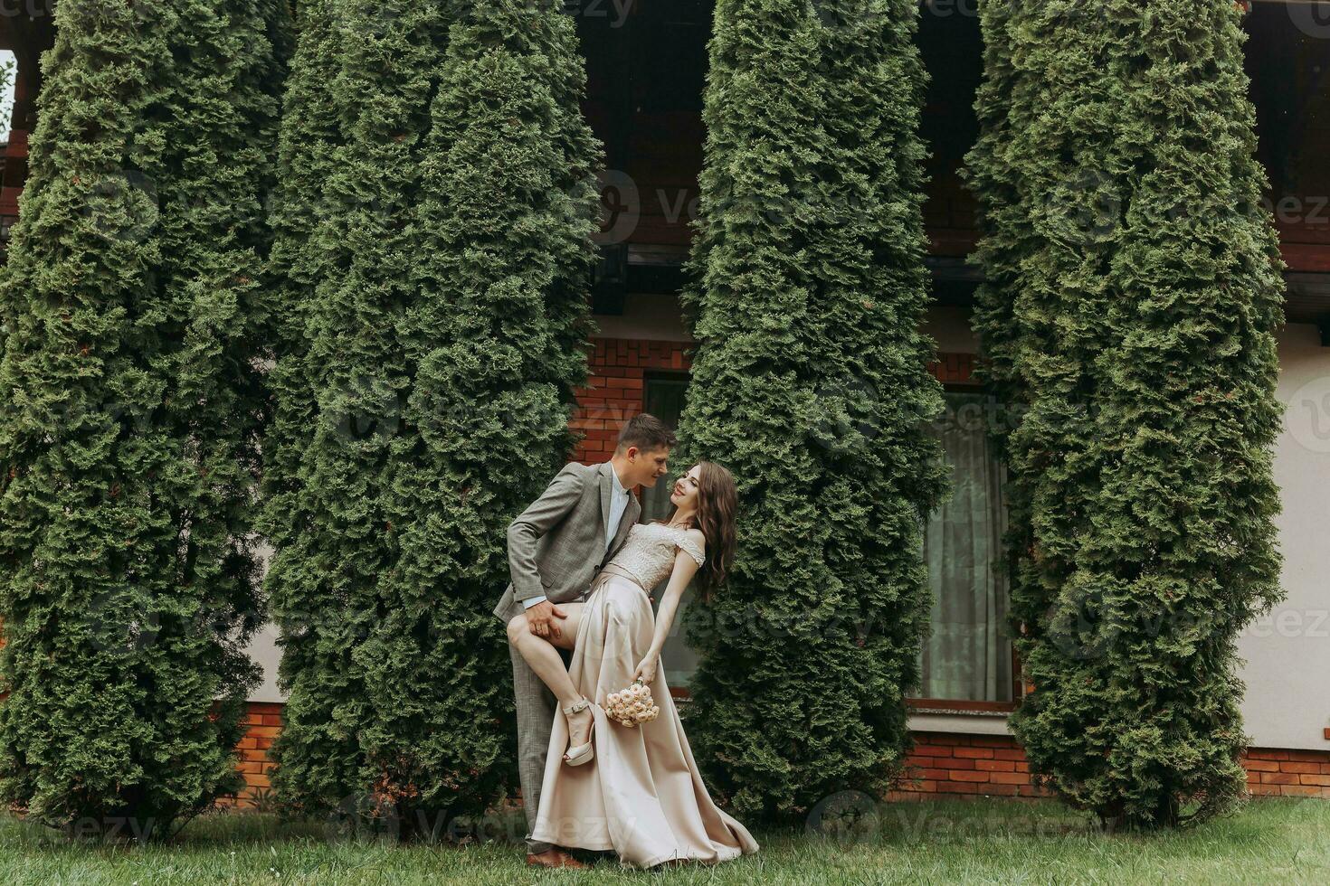 Groom and bride in the park in nature, green thuja trees on the background. Kiss. Photo portrait. Wedding couple