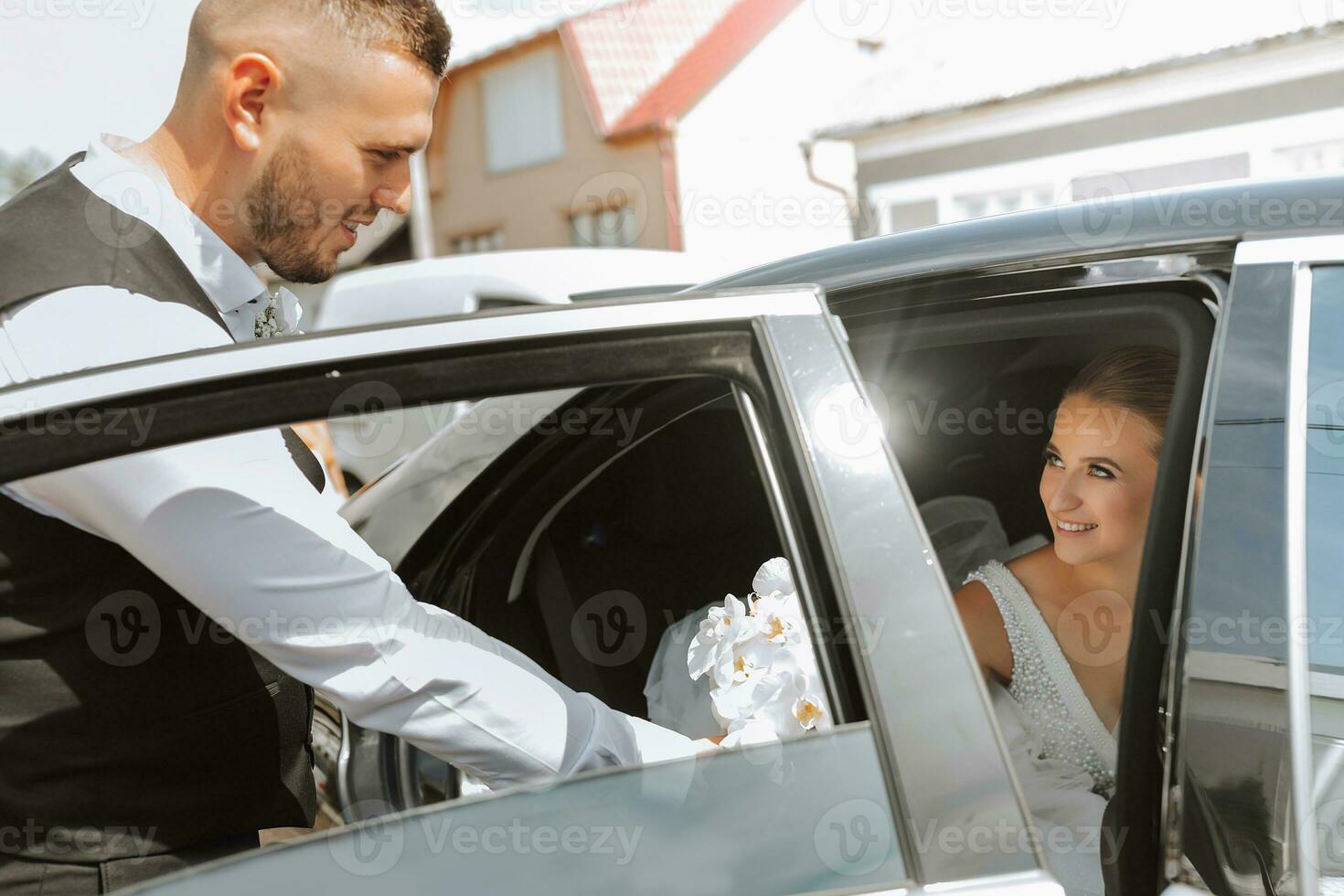 A blonde bride, smiling sincerely, sits in a black car on her wedding day and gives her hand to the groom, looking at him. Portrait of the bride. Beautiful makeup and hair. A luxurious white dress photo