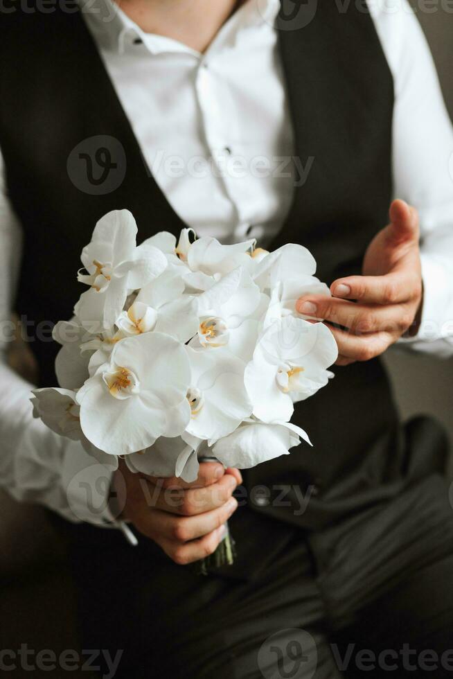 A stylish groom dressed in a classic suit holds a bouquet of orchid flowers in his hands. Groom's morning. The groom is getting ready in the morning before the wedding ceremony photo