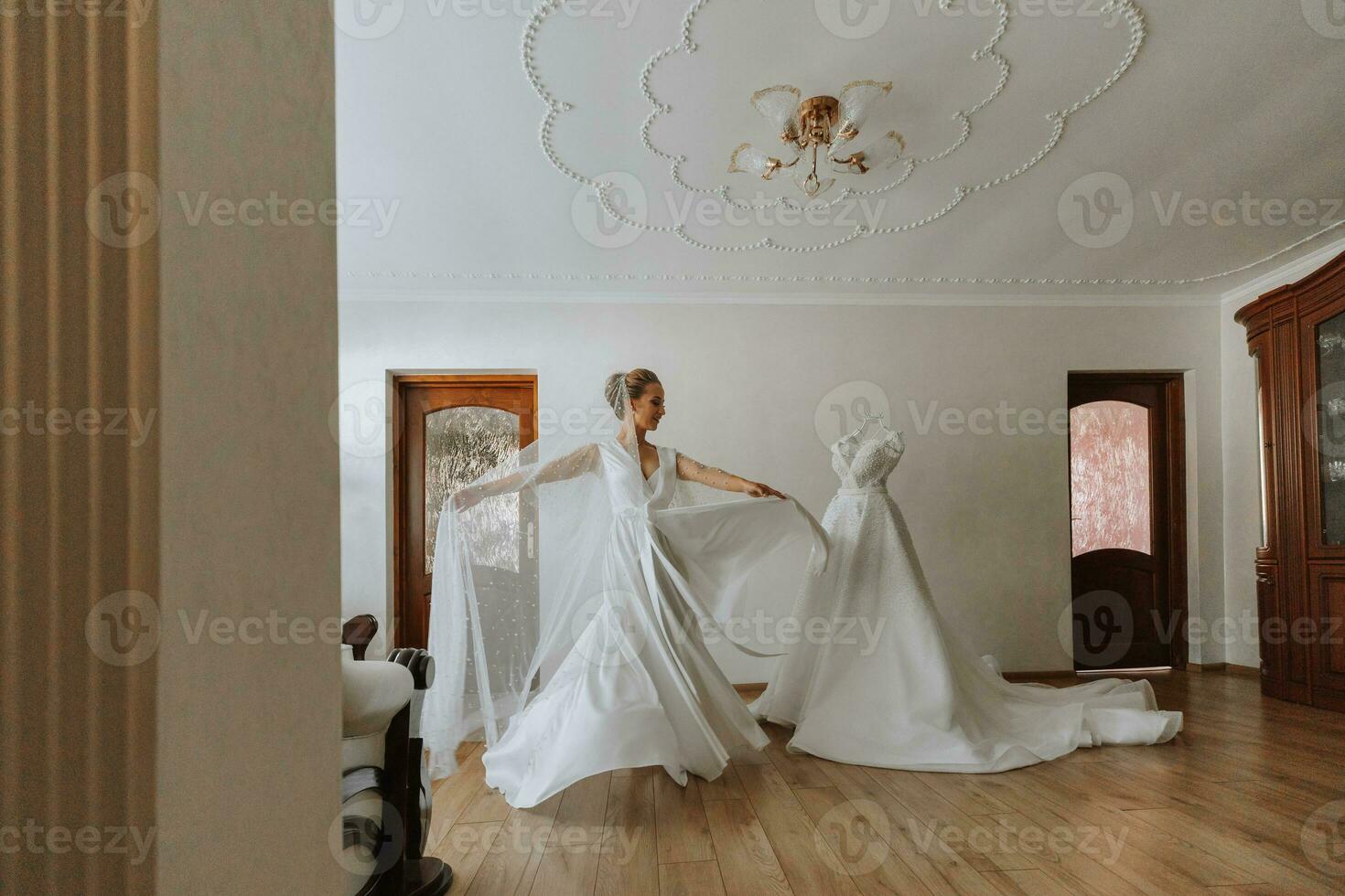 A luxurious bride with a classic hairstyle, wearing a long robe and a long veil, is circling her wedding dress on a mannequin in her room. The bride on a white background photo