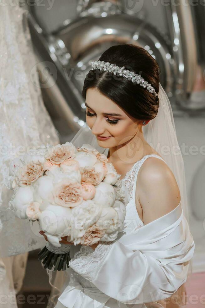 A brunette bride in a white robe and veil poses by the bed with a bouquet of peonies. Diadem. Wedding concept. Morning of the bride. Classic hair and makeup photo