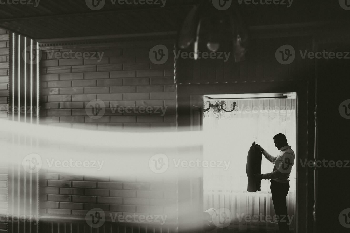 A man in a white shirt holds his gray jacket against the background of the window. Side view. Wide angle black and white photo. Men's style. Fashion. Business photo