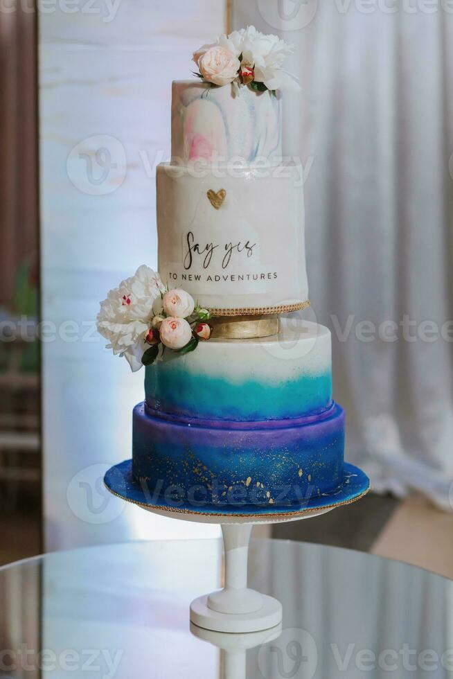 A four-tier white and blue wedding cake decorated with peonies on a mirrored surface in the wedding hall. photo