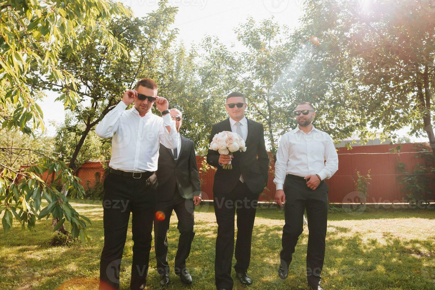 The groom in a black suit and glasses and his stylish friends wearing white shirts and black pants and glasses are standing in the backyard in the garden. The groom is holding a bouquet. photo